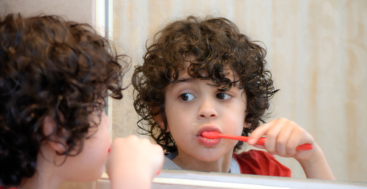 Young_boy_with_dark_curly_hair_looking_in_the_mirror_brushing_his_teeth_with_an_orange_toothbrush