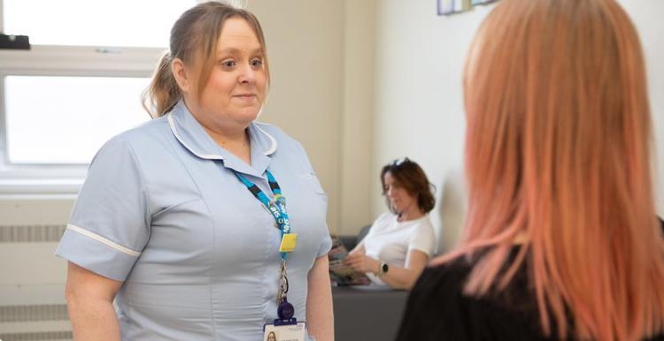 female_nurse_wearing_blue_scrubs_talking_to_a_woman_with_red_hair_with_a_woman_sitting_reading_in_the_background