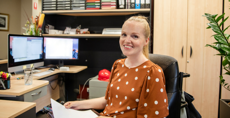 Young_woman_sitting_at_a_desk_smiling.She_is_wearing_a_brown_top_with_white_spots_and_has_a_blonde_ponytail