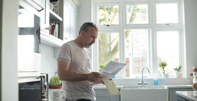 A_middle_aged_man_wearing_a_white_t-shirt_with_grey_hair_is_leaning_against_a_kitchen_worktop_looking_at_a_letter