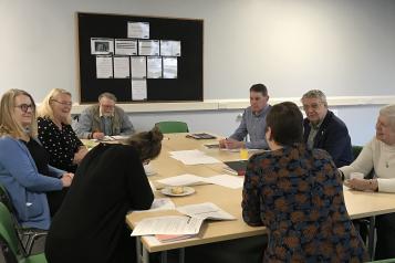 8 people. 5 female and 3 male sat around a table with papers on, in a meeting room