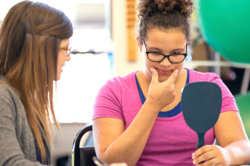 Young_woman_looking_into_a_mirror_practising_her_speech_therapy_whilst_her_female_tutor_helps
