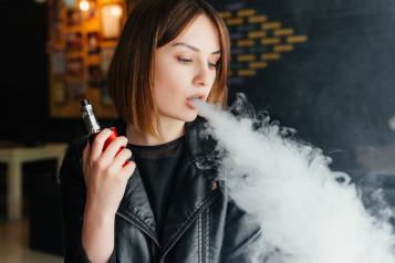 Woman_with_shoulder_length_brown_hair_wearing_a_black_biker_jacket_blowing_out_smoke_from_a_vape_device
