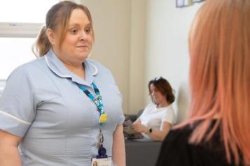 female_nurse_wearing_blue_scrubs_talking_to_a_woman_with_red_hair_with_a_woman_sitting_reading_in_the_background