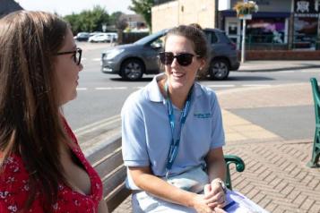 A_woman_wearing_a_red_top_and_brown_hair_sitting_on_a_bench_talking_to_person_with_blue_tshirt_and_sunglasses