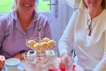 Two_women_smiling_sitting_at_a_small_table_drinking_tea_and_eating_scones