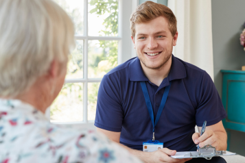 man_wearing_a_navy_blue_polo_shirt_sitting_down_smiling_with_a_clipboard_talking_to_an_older_woman