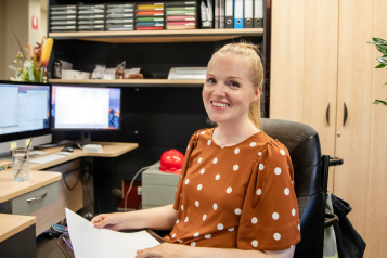 Young_woman_sitting_at_a_desk_smiling.She_is_wearing_a_brown_top_with_white_spots_and_has_a_blonde_ponytail