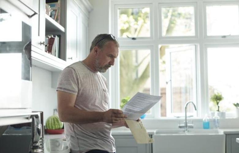 A_middle_aged_man_wearing_a_white_t-shirt_with_grey_hair_is_leaning_against_a_kitchen_worktop_looking_at_a_letter